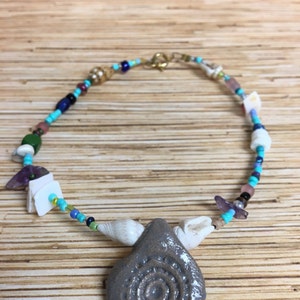 May include: A handmade necklace featuring a gray pendant with a spiral design, seashells, and colorful beads. The necklace includes turquoise, blue, and white beads, with a gold-colored clasp. The pendant is teardrop-shaped.