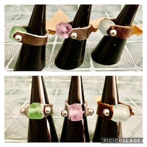 May include: A collection of handcrafted rings featuring various colored gemstones, including green, pink, and blue, set in brown leather bands. Each ring has silver-colored metal accents and is displayed on a black ring holder.