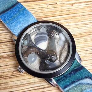 May include: A unique wristwatch with a black bezel and a blue and white patterned band. The watch face features a miniature beach scene with sand, shells, and shark teeth. The band is approximately 0.75 inches wide.