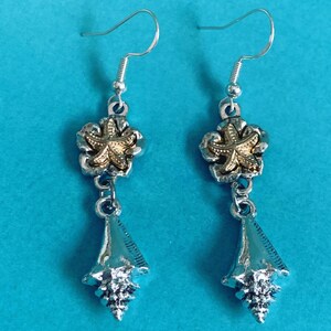May include: A pair of silver-toned earrings with a beach theme. Each earring features a gold-colored starfish charm set within a silver-toned flower, with a dangling silver-toned seashell pendant. The earrings hang from silver-colored fish hook ear wires.