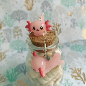 May include: A pair of pink axolotl earrings. The earrings feature a cute axolotl design with pink and red accents. The earrings are displayed on a cork and glass jar filled with seashells.
