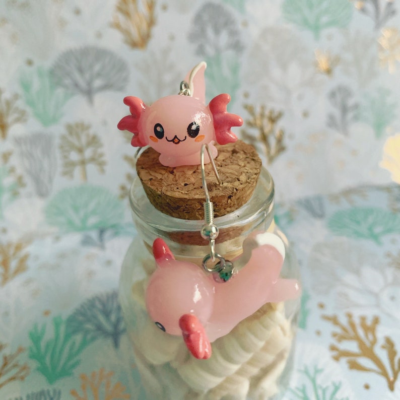May include: A pair of pink axolotl earrings. The earrings feature a cute axolotl design with pink and red accents. The earrings are displayed on a cork and glass jar filled with seashells.