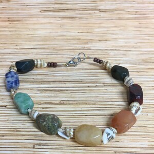 May include: A handmade bracelet featuring a variety of colorful gemstones and small seashells. The bracelet includes stones in shades of blue, green, yellow, orange, and brown, interspersed with tiny seashells. It is strung on a thin, brown cord and secured with a silver clasp.