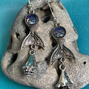 May include: A pair of silver-toned mermaid earrings. Each earring features a round, iridescent scale-like top, a mermaid tail, and a dangling seashell charm. The earrings are displayed on a gray stone against a blue background.