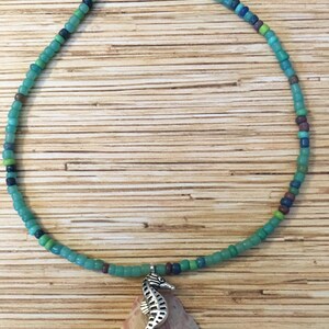 May include: A beaded necklace with a seahorse pendant. The necklace features turquoise, green, and blue beads. A silver seahorse charm hangs from the necklace, resting on a pink and purple seashell.
