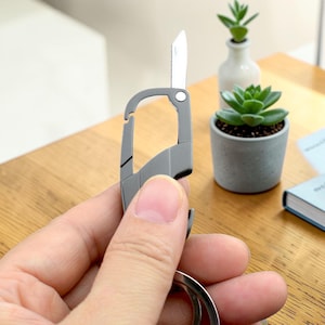 May include: A matte gray metal multi-tool carabiner featuring an integrated folding blade and a bottle opener. The tool has a sleek, angular design with a spring-loaded gate mechanism and a circular attachment ring at the base for keys.