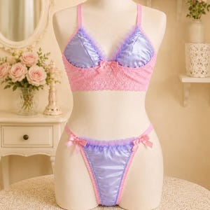 Pode incluir: Um conjunto de lingerie de duas peças composto por um bralette de cetim lavanda e uma tanga de corte alto a combinar. O bralette tem acabamento em renda rosa, alças finas e um pequeno laço central. A tanga inclui bordas de renda rosa nas laterais.