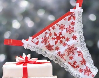 May include: White and red thong underwear with a snowflake pattern and white lace trim. The straps are red with white bows. A stack of white gift boxes with red ribbons is in the foreground.