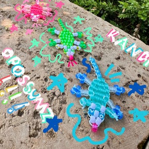 Könnte beinhalten: Drei Perleidechsenfiguren in Rosa, Grün und Blau mit weißen und farbigen Akzenten werden auf einem Felsen gezeigt. Das Wort "Kandi" ist in Pastellfarben geschrieben, zusammen mit dem Wort "Grosssumz" und anderen bunten Kritzeleien.