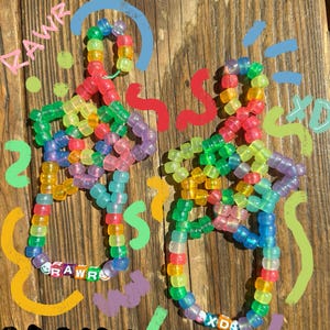 Könnte beinhalten: Zwei bunte Perlenarmbänder mit den Wörtern "RAWR" und "XD" aus Perlen. Die Armbänder sind aus verschiedenen Farben gefertigt und liegen auf einer Holzoberfläche. Die Worte "GLOWS IN THE DARK" sind oberhalb und unterhalb der Armbänder geschrieben.