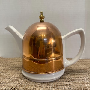 May include: A copper and white teapot with a rounded copper lid and a small gold-colored finial. The teapot has a white handle and spout. The base is white, and the teapot sits on a woven mat.