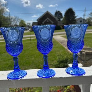 May include: Three cobalt blue goblets with a detailed design. Each goblet features a textured diamond pattern on the bowl and stem, with a medallion of a woman's profile. The goblets are displayed outdoors.