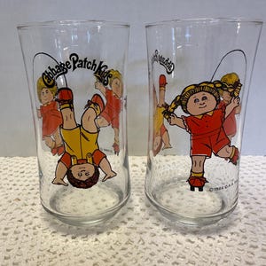 May include: Two clear glass tumblers featuring Cabbage Patch Kids graphics. One tumbler shows a child upside down, the other shows a child jumping rope. The text "Cabbage Patch Kids" is printed on the glasses.