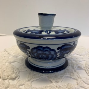 May include: A small, white ceramic jar with a blue and white floral design. The jar has a lid with a knob on top. The jar is sitting on a white crocheted doily.