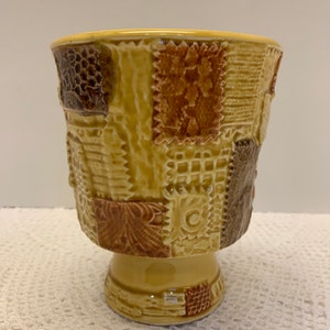 May include: A yellow ceramic planter with a pedestal base. The planter is decorated with a patchwork design in shades of brown and yellow.