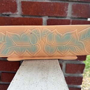 May include: A rectangular, peach-colored ceramic planter with a raised leaf design in teal. The planter sits on a small base and is displayed against a red brick wall and a white surface.