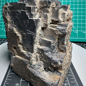 May include: A large, dark gray galena mineral specimen with a cubic crystalline structure. The surface features sharp, stepped geometric formations and metallic luster, interspersed with rough, light-colored mineral matrix deposits.