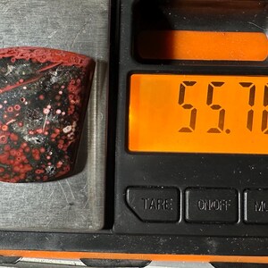Op de afbeelding: Een gepolijste edelsteen met een rood en zwart patroon, weergegeven op een weegschaal. De weegschaal toont een gewicht van 55,70 karaat. De edelsteen heeft een unieke vorm en een glanzende afwerking, met rode en witte vlekken.