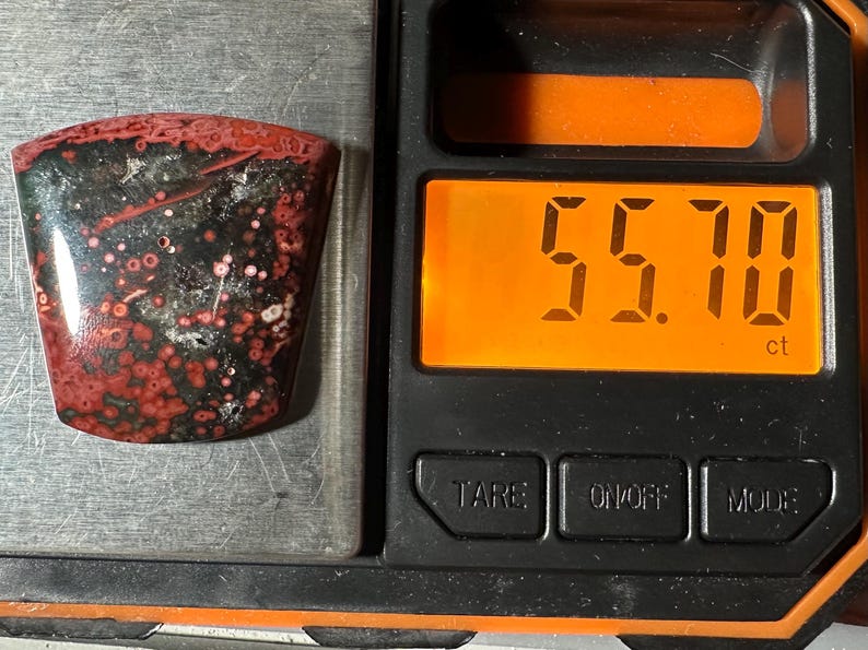 Op de afbeelding: Een gepolijste edelsteen met een rood en zwart patroon, weergegeven op een weegschaal. De weegschaal toont een gewicht van 55,70 karaat. De edelsteen heeft een unieke vorm en een glanzende afwerking, met rode en witte vlekken.