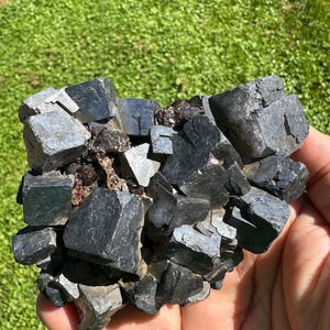 May include: A cluster of metallic, dark gray galena crystals with cubic formations. The mineral specimen features sharp, geometric edges and facets with a reflective, lead-like luster, interspersed with small, dark reddish-brown inclusions.