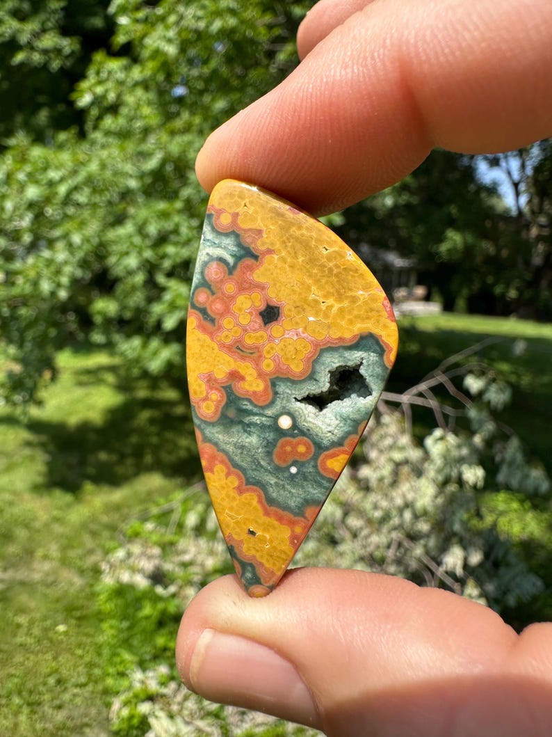 Puede incluir: Un cabuj&oacute;n de jaspe oce&aacute;nico pulido, de forma triangular, de color verde, naranja y rojo con una mancha blanca en el centro.