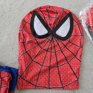 Puede incluir: M&aacute;scara roja de Spider-Man con un patr&oacute;n de telara&ntilde;a negra y aberturas para los ojos blancas. La m&aacute;scara es parte de un disfraz, con una manga roja y azul a juego visible. La m&aacute;scara est&aacute; hecha de un material flexible.