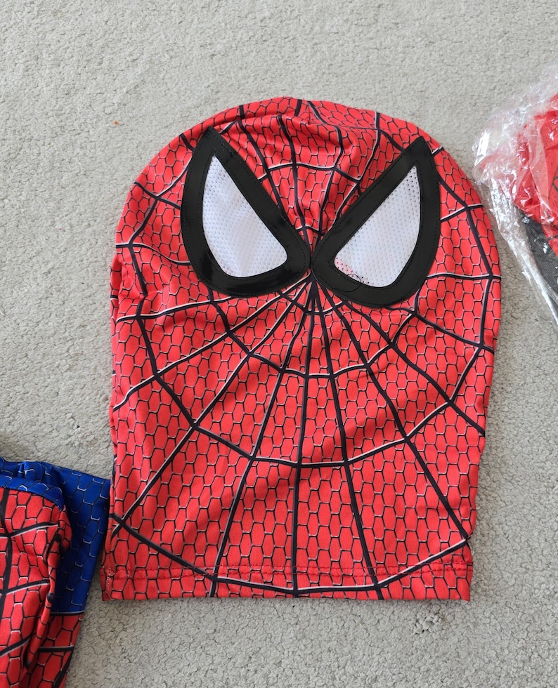 Puede incluir: M&aacute;scara roja de Spider-Man con un patr&oacute;n de telara&ntilde;a negra y aberturas para los ojos blancas. La m&aacute;scara es parte de un disfraz, con una manga roja y azul a juego visible. La m&aacute;scara est&aacute; hecha de un material flexible.