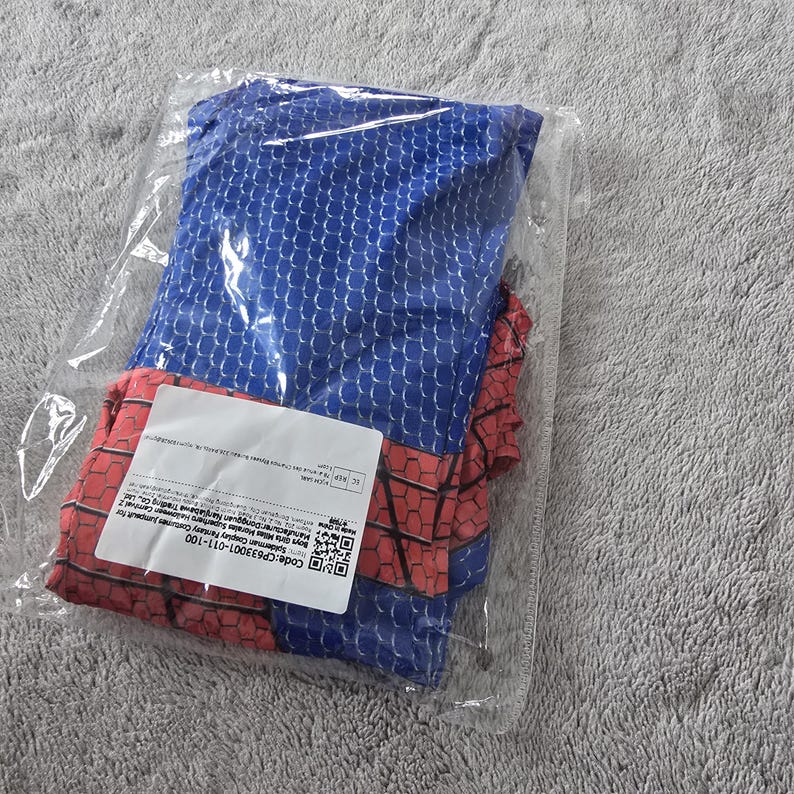 Puede incluir: Un disfraz de Spiderman azul y rojo, doblado y empaquetado en una bolsa de pl&aacute;stico transparente. El disfraz presenta una parte superior azul con un patr&oacute;n de escamas y secciones rojas. Una etiqueta blanca con texto y un c&oacute;digo de barras es visible.
