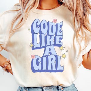 May include: Cream-colored t-shirt with the text "CODE LIKE A GIRL" in a retro blue font. The design includes stars and flowers. A casual, comfortable tee for those who love coding.