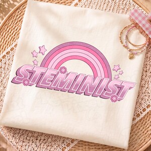 Puede incluir: Una camiseta de color crema con un gráfico de un arcoíris rosa y morado con el texto STEMINIST en una fuente retro llamativa. El diseño incluye pequeñas estrellas y flores rosas, acompañado de una pulsera de cuentas dorada con un dije de corazón.