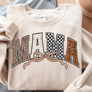 May include: Cream-colored sweatshirt with the word "MAMA" in a bold, decorative font. The letters feature various patterns, including leopard print, gingham, and a black and white checkerboard design. A tan bow is beneath the word.