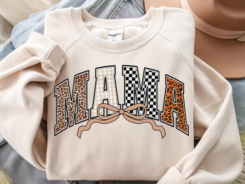 May include: Cream-colored sweatshirt with the word "MAMA" in a bold, decorative font. The letters feature various patterns, including leopard print, gingham, and a black and white checkerboard design. A tan bow is beneath the word.