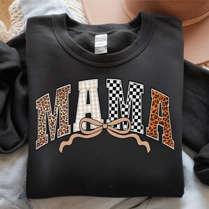 May include: Black sweatshirt with the word "MAMA" in a bold, arched design. The letters feature a mix of patterns: leopard print, gingham, and a black and white checkerboard. A tan bow is beneath the word. The sweatshirt is folded on a light-colored surface.