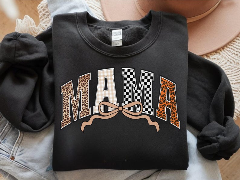 May include: Black sweatshirt with the word "MAMA" in a bold, arched design. The letters feature a mix of patterns: leopard print, gingham, and a black and white checkerboard. A tan bow is beneath the word. The sweatshirt is folded on a light-colored surface.