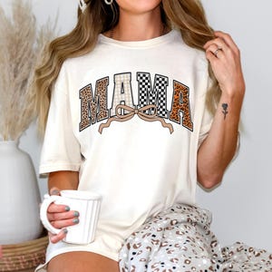 May include: Cream-colored t-shirt with the word "MAMA" in a collegiate style. The letters feature leopard print, checkered, and plaid patterns. A tan bow is beneath the word. The shirt is part of a casual wear collection.
