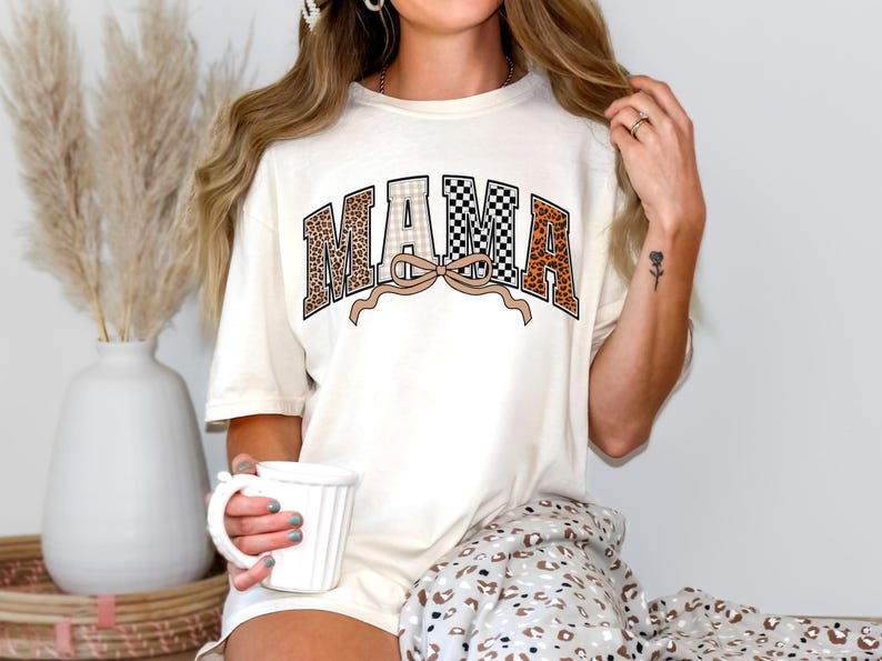 May include: Cream-colored t-shirt with the word "MAMA" in a collegiate style. The letters feature leopard print, checkered, and plaid patterns. A tan bow is beneath the word. The shirt is part of a casual wear collection.