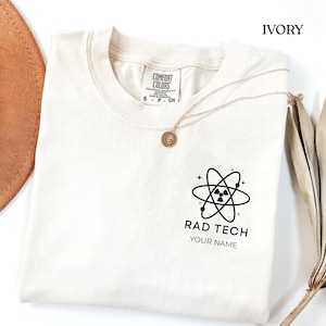 May include: Ivory-coloured t-shirt featuring a black atom graphic and the text "RAD TECH" and "YOUR NAME". The shirt has a crew neck and short sleeves. A gold necklace with a pendant is draped across the shirt.
