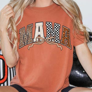 May include: Rust-colored t-shirt with the word "MAMA" in a decorative design. The letters feature leopard print, checkered, and plaid patterns, with a tan bow beneath the word. The shirt is a casual, comfortable style.
