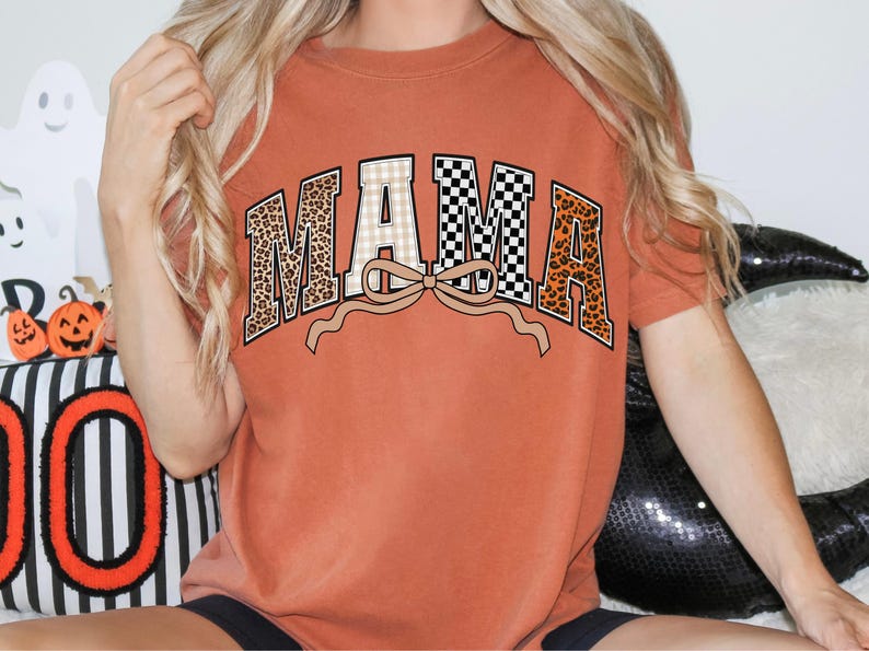 May include: Rust-colored t-shirt with the word "MAMA" in a decorative design. The letters feature leopard print, checkered, and plaid patterns, with a tan bow beneath the word. The shirt is a casual, comfortable style.
