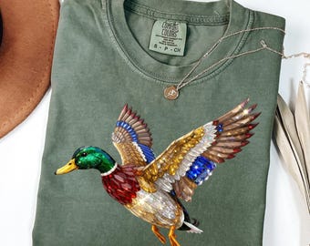 Può includere: Una t-shirt girocollo verde oliva con una grafica di un germano reale resa in uno stile sfaccettato color gemma con vivaci colori rosso, blu, oro e bianco. La maglietta presenta un'etichetta del marchio Comfort Colors all'interno del colletto.