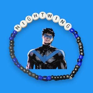 Puede incluir: Una pulsera de cuentas con la palabra "NIGHTWING" escrita con cuentas de letras blancas. La pulsera presenta cuentas negras, grises y azules. Una ilustración de cómic de Nightwing está en el fondo, sobre un fondo azul.