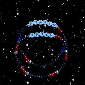 Op de afbeelding: Drie armbanden met kralen met de namen "ANAKIN" en "VADER" gespeld in witte letterkralen. De armbanden zijn zwart met rode en blauwe accenten. De achtergrond is een zwarte ruimtescène met witte sterren.