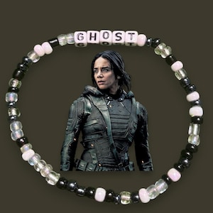 Può includere: Un braccialetto di perline con la parola "GHOST" scritta con perline bianche. Il braccialetto presenta un mix di perline nere, trasparenti e rosa. Lo sfondo include un'immagine di una persona in abito nero.