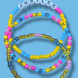 May include: Colorful beaded bracelets with the word "JUBILEE" spelled out in white letter beads. The bracelets feature a mix of yellow, pink, blue, and clear beads. The background is a solid light blue.