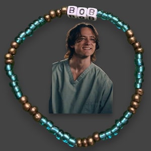 May include: A beaded bracelet with the name "BOB" spelled out in white letter beads. The bracelet features alternating teal and bronze-colored beads. A photo of a smiling person in a teal shirt is in the center.