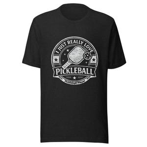 Può includere: T-shirt nera con grafica bianca. Il design presenta una racchetta da pickleball, una palla e stelle all'interno di un emblema circolare. Il testo recita "I Just Really Love Pickleball" e "Pickleball Player".
