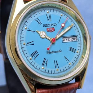 May include: A gold-tone Seiko 5 automatic wristwatch with a light blue face and brown leather band. The watch face features Roman numerals, a day-date display, and red second hand. The watch is displayed on a black stand.
