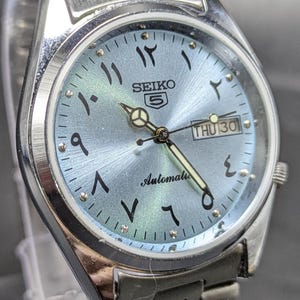 May include: A silver-tone Seiko 5 automatic wristwatch featuring a light blue sunray dial with black Eastern Arabic numerals, luminous hands, and a day-date window at the three o'clock position. The watch includes a stainless steel link bracelet.