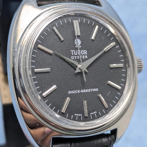 May include: A vintage Tudor Oyster watch with a black dial and silver hands and markers. The watch has a stainless steel case and a black leather strap. The dial reads "Tudor Oyster" and "Shock-Resisting".