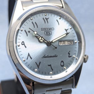 May include: A silver Seiko 5 automatic wristwatch with a light blue dial. The watch features Arabic numerals, a day-date display, and a silver bracelet. The watch hands and hour markers are silver. The watch face reads "Automatic".